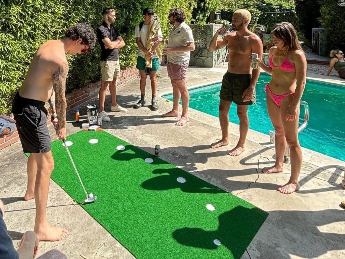 A group of people playing miniature golf outside near a swimming pool on a sunny day. One person is preparing to putt while others watch.