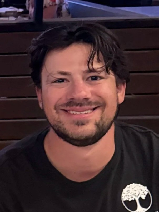 A man with dark, wavy hair and a beard smiling at the camera. He is wearing a black T-shirt with a white tree logo on the chest. The background has dark wooden panels.