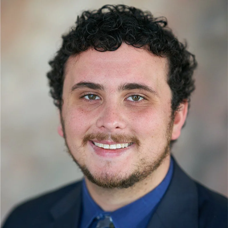 Portrait of a young man with curly dark hair, light skin, and facial hair, wearing a dark suit and blue shirt, smiling at the camera.