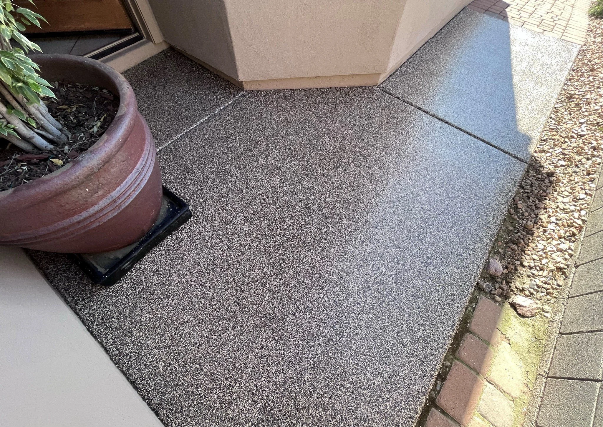 Close-up of a textured concrete porch with a large potted plant on a black tray, adjacent to a beige wall and a brick pathway.
