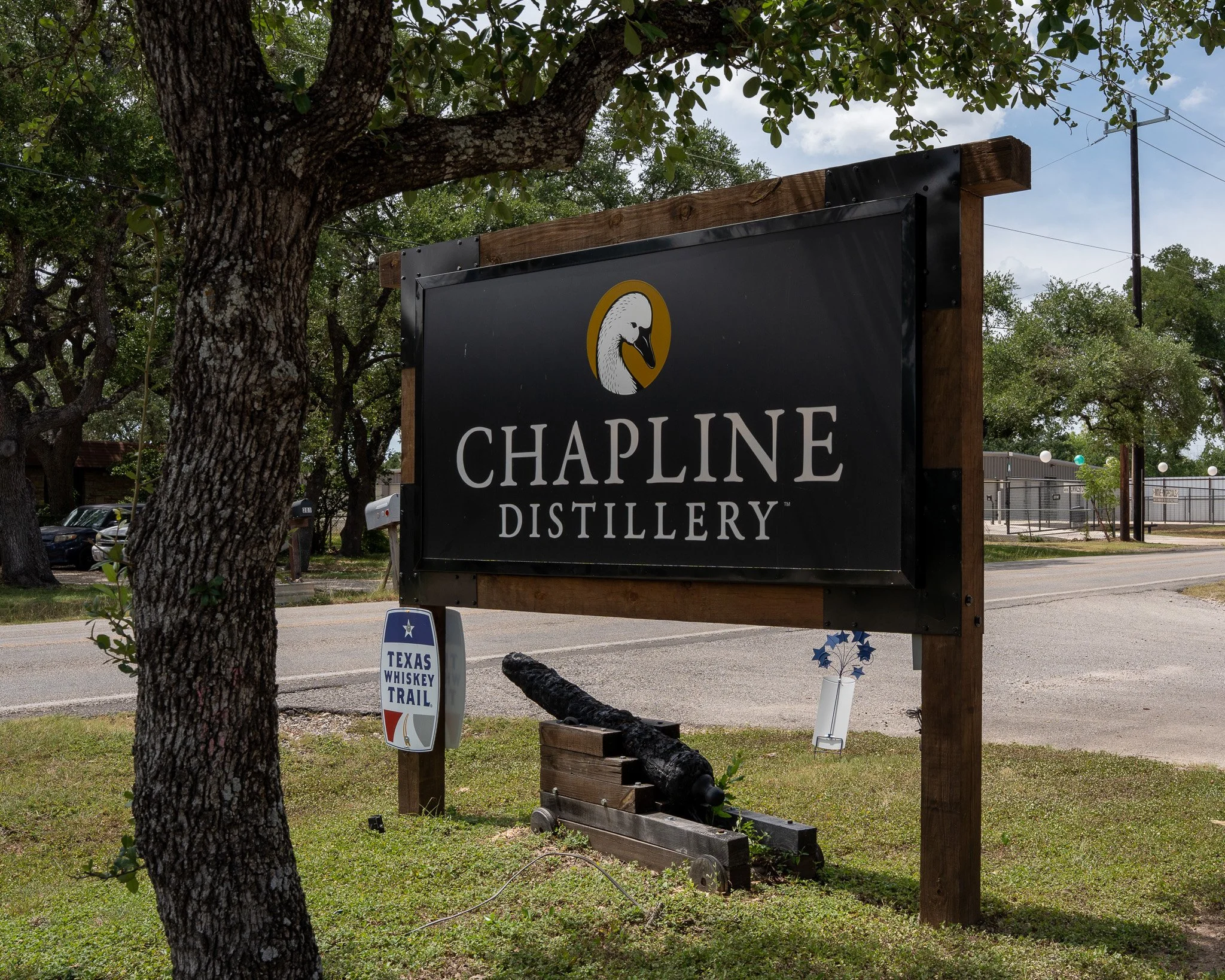 A sign with the words Chapline Distillery on the side of a road with a small historical cannon positioned beneath it. There is also a Texas Whiskey Trail sign
