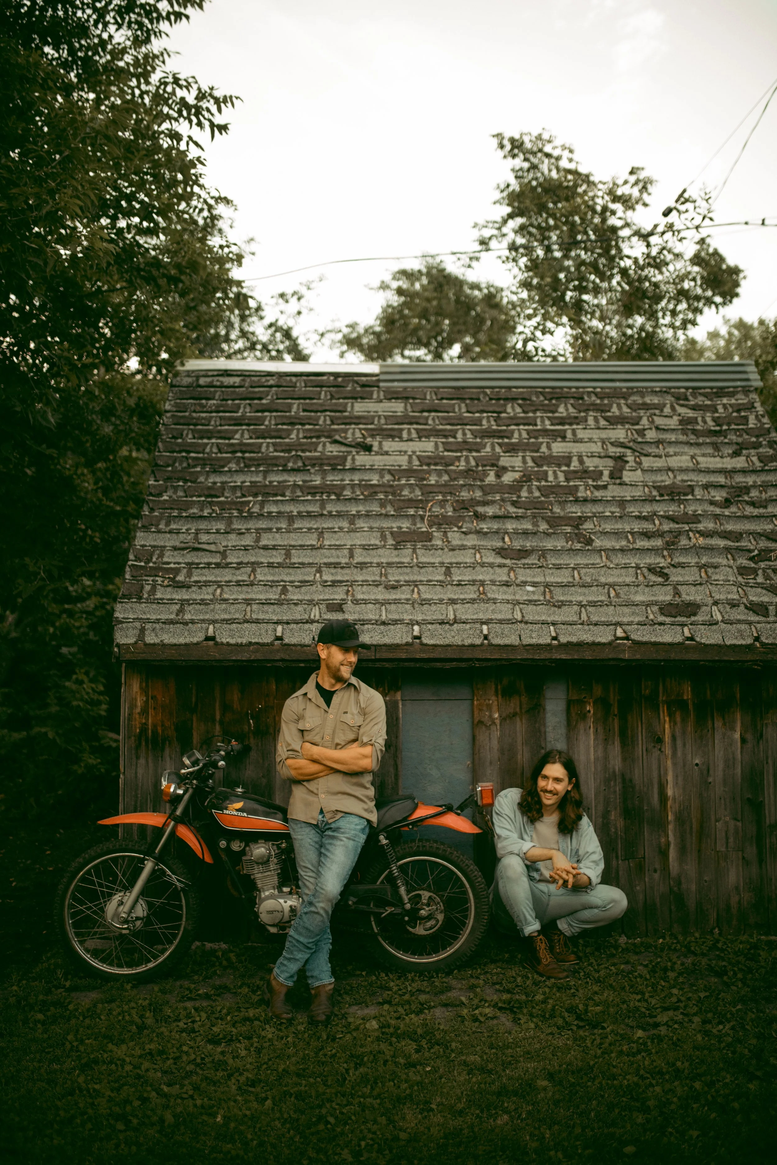 Two men outdoors, one leaning against a motorcycle and the other sitting on the grass, smiling. They are in front of a wooden shed with a shingled roof and trees in the background.