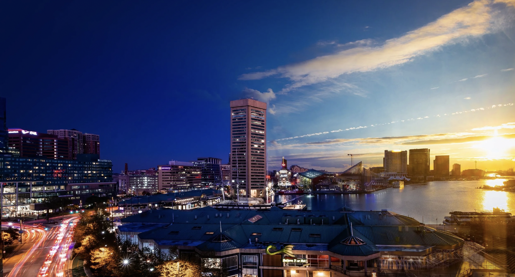 City skyline at sunset with tall buildings, a river, and cloudy sky.
