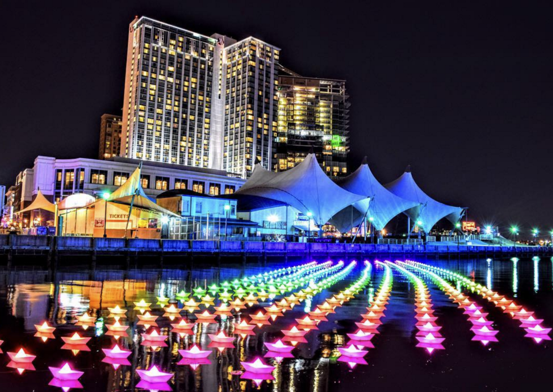 Night view of a city waterfront with colorful star-shaped lights floating on the water, modern high-rise buildings with illuminated windows, and a large tent-like structure on the pier.