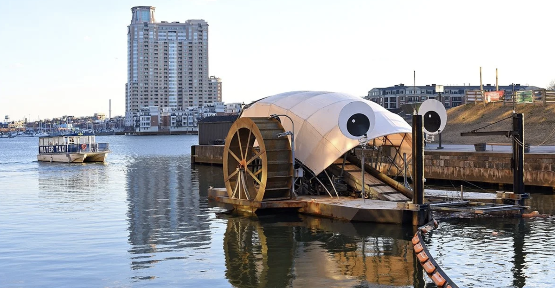 A floating structure with large cartoonish eyes on a body of water, resembling a whimsical watercraft or attraction, with a city skyline and a water taxi in the background.