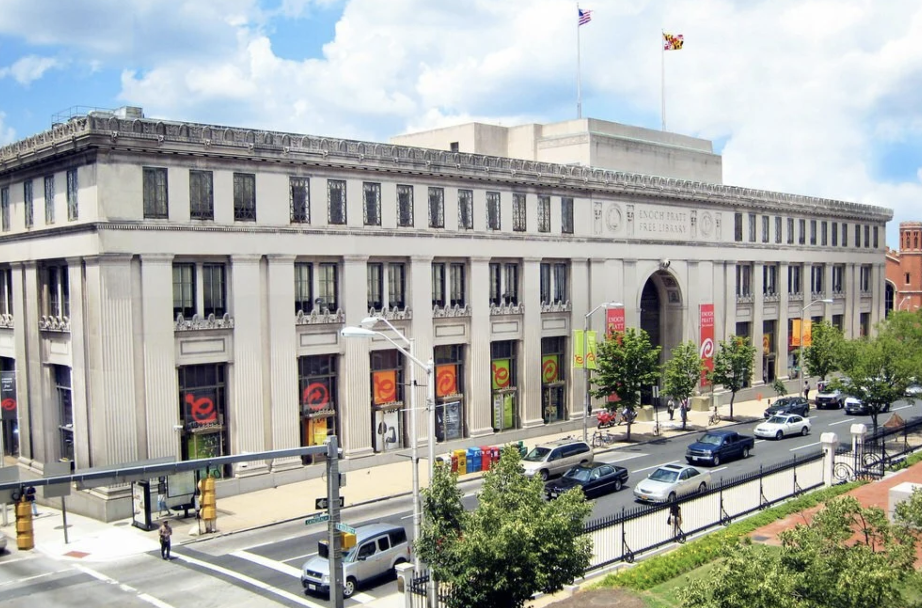 A large classic-style library building with a sign that reads 'ENOCH PRATT FREE LIBRARY' at the top center. Several flags are flying on top, and the street in front has parked cars, pedestrians, and trees.