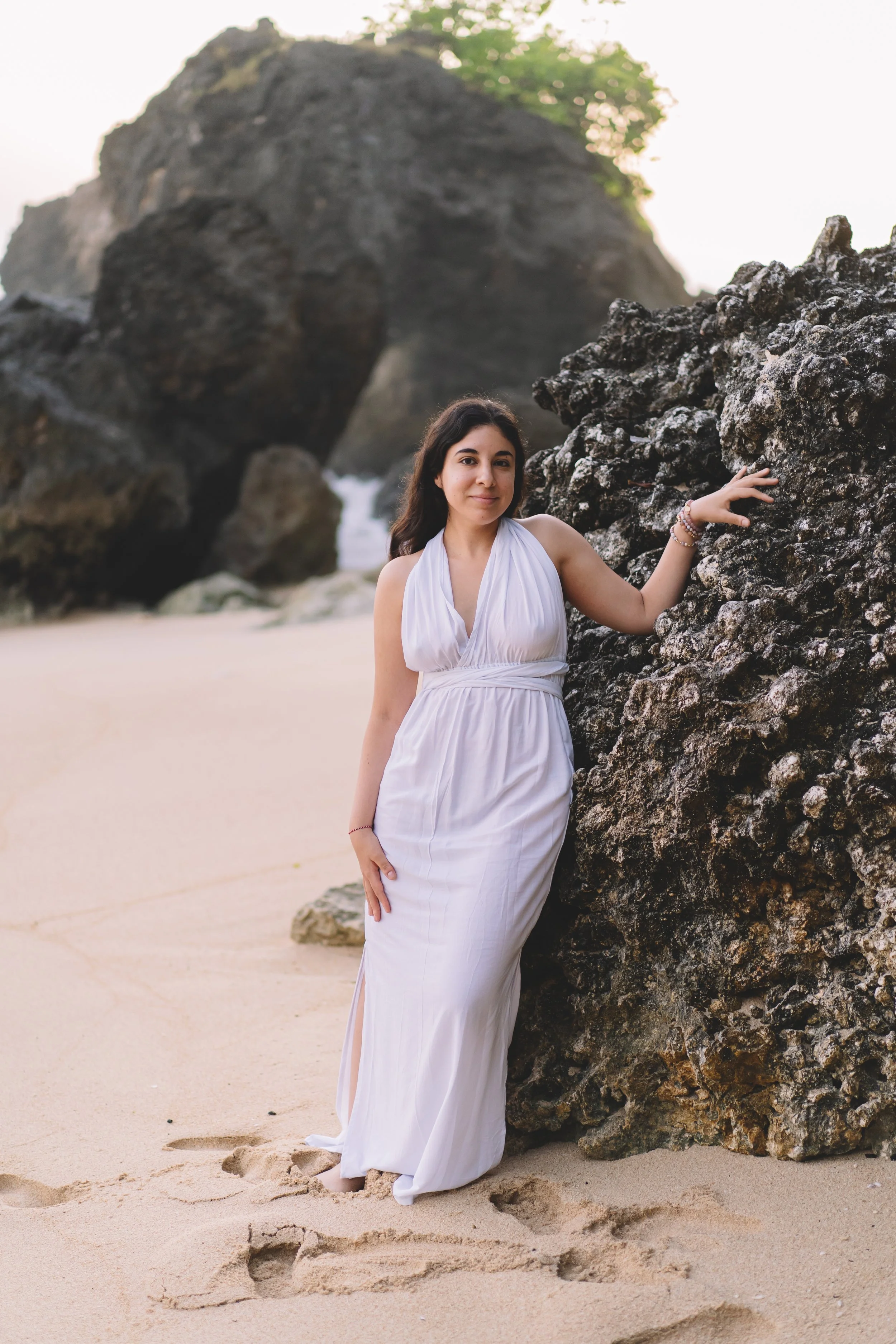 A woman in a white dress standing on a sandy beach, leaning against a large rocky formation with another big rock in the background and greenery at the top.
