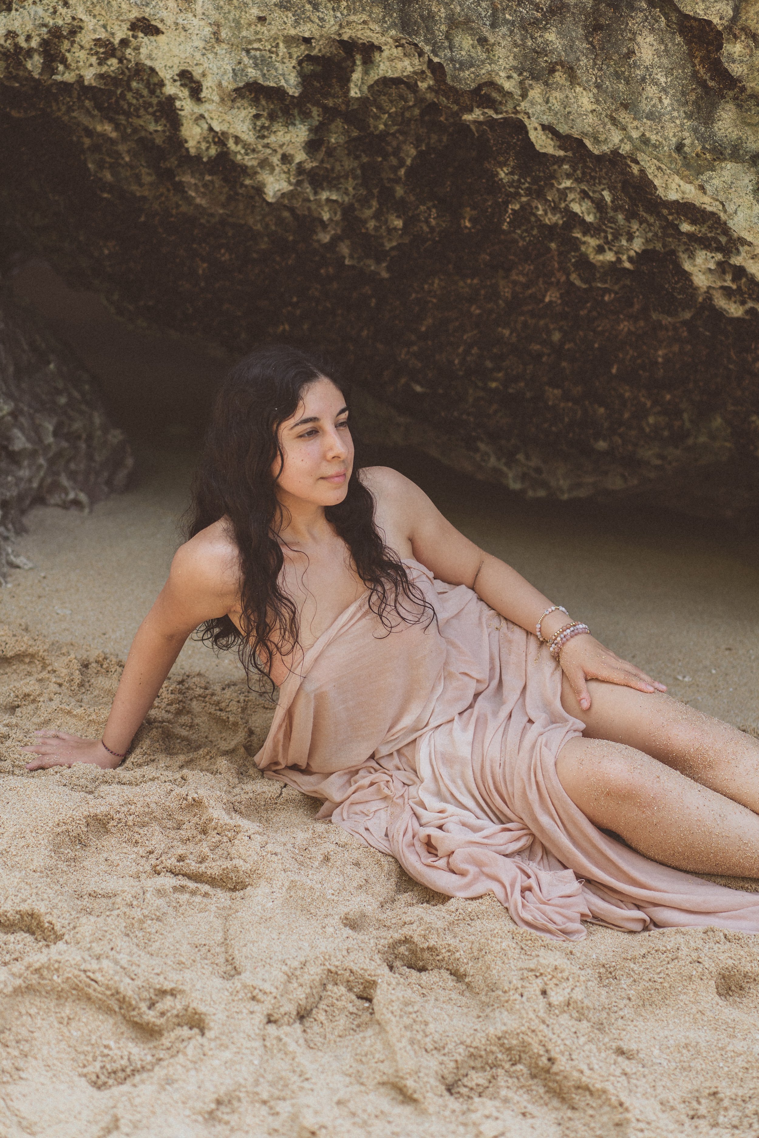 A woman in a pink dress sitting on sandy ground beneath a large rock formation
