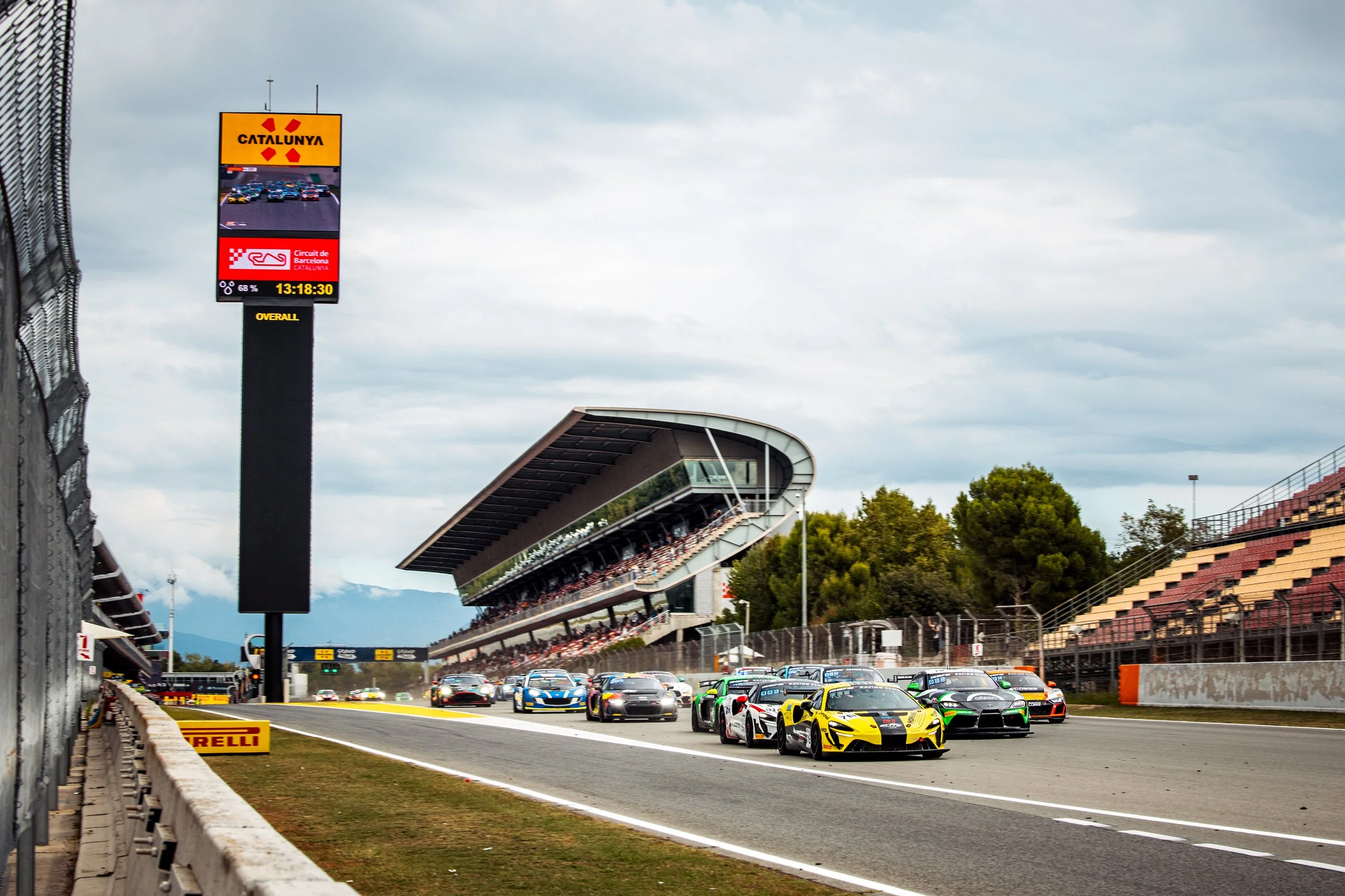 Race cars on a race track at Circuit de Barcelona-Catalunya in Spain, with a digital scoreboard showing the race details and a grandstand in the background.