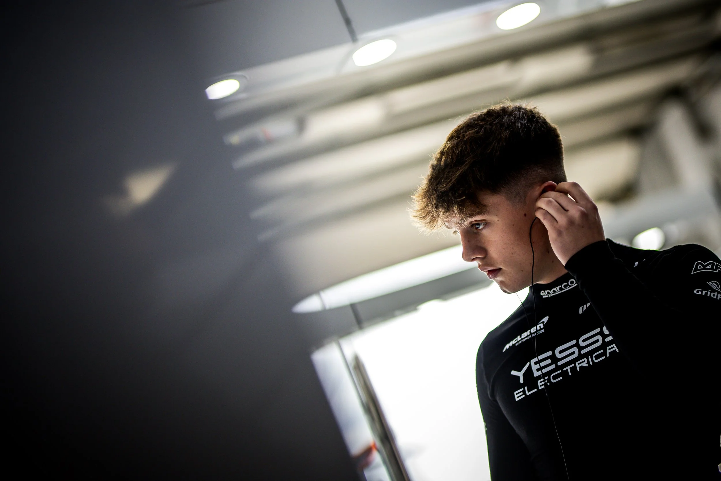 Close-up of a young man with brown hair wearing a black racing suit with 'YESS! ELECTRICA' written on it, adjusting his earbud in a modern indoor setting.