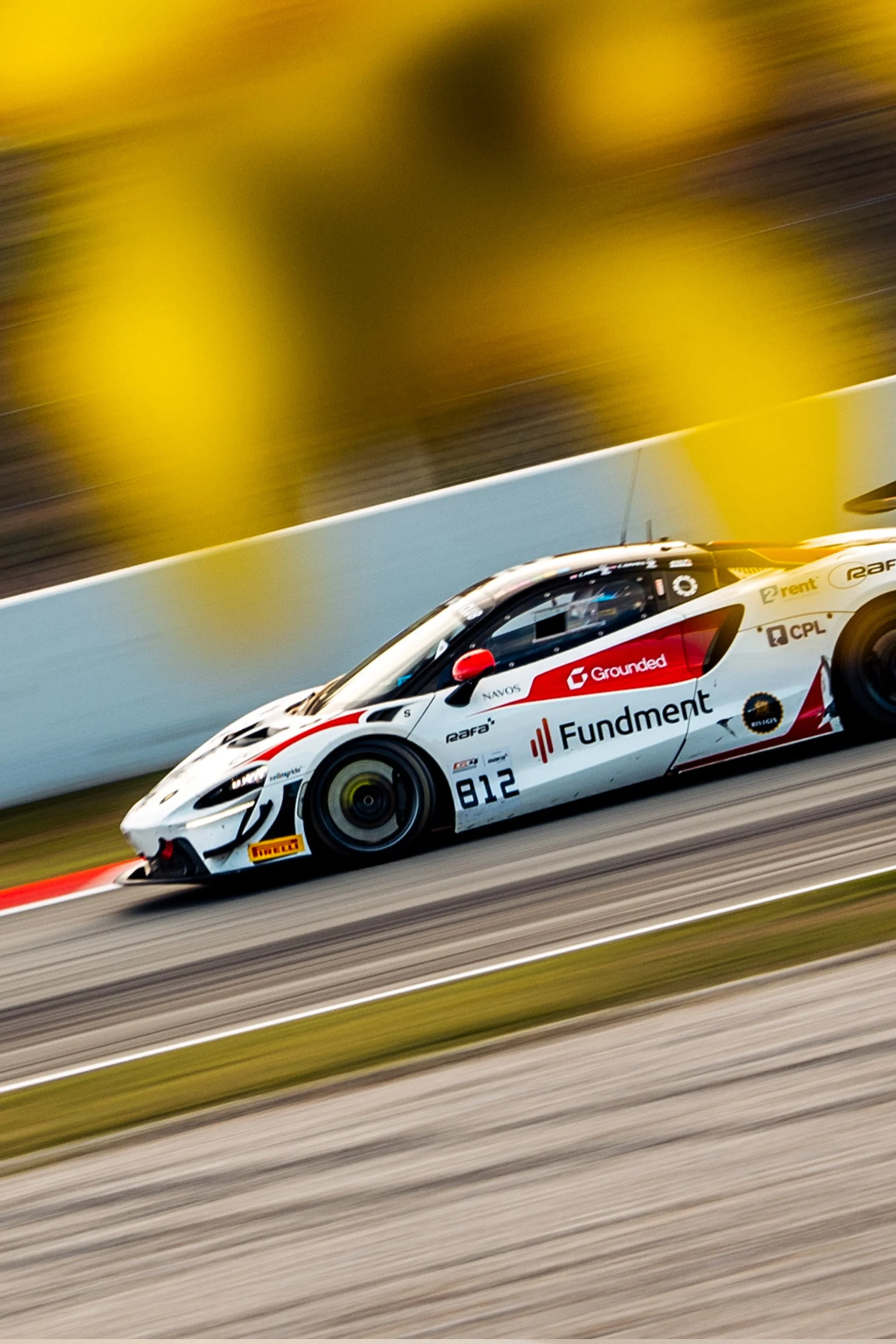 A race car speeding on a race track with motion blur in the background. The car is white with red and black accents, and has various sponsor logos including 'Fundment', 'Grounded', and 'Pirelli'. The number 812 is visible on the side of the car.