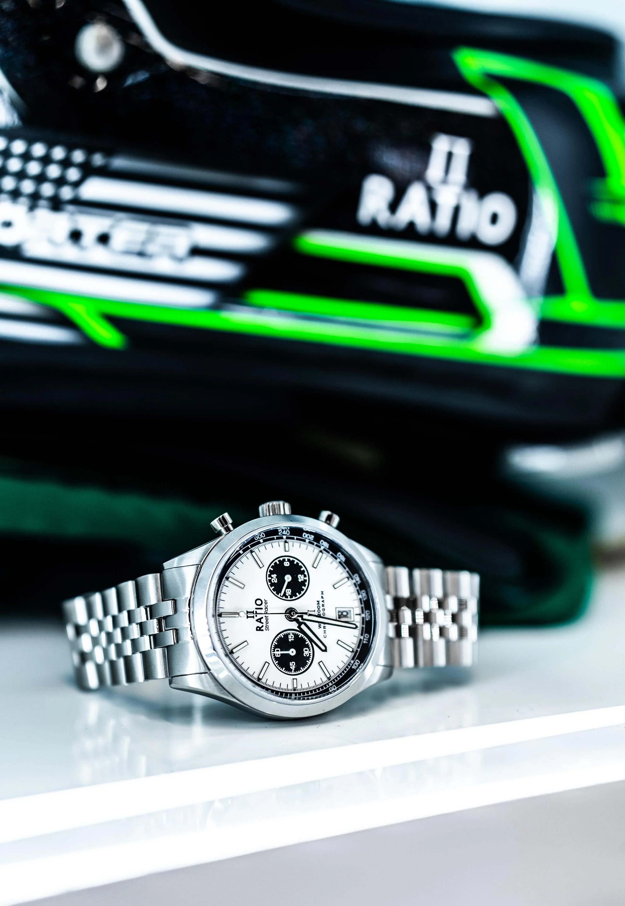 A silver wristwatch with a white dial, black subdials, and a metal bracelet, placed on a white surface. In the background, a blurred black object with green accents and the word "RATIO" is visible.