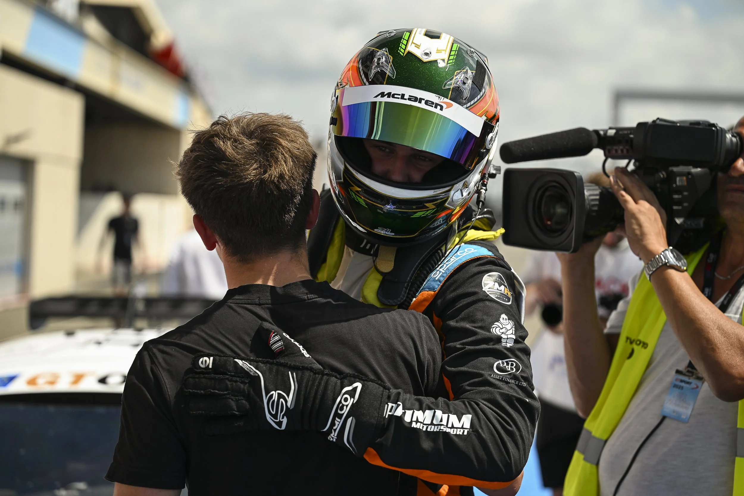 A race car driver in racing gear, including a helmet, embraces a team member in a paddock area. A cameraman interviews them nearby, with other people and racing infrastructure in the background.