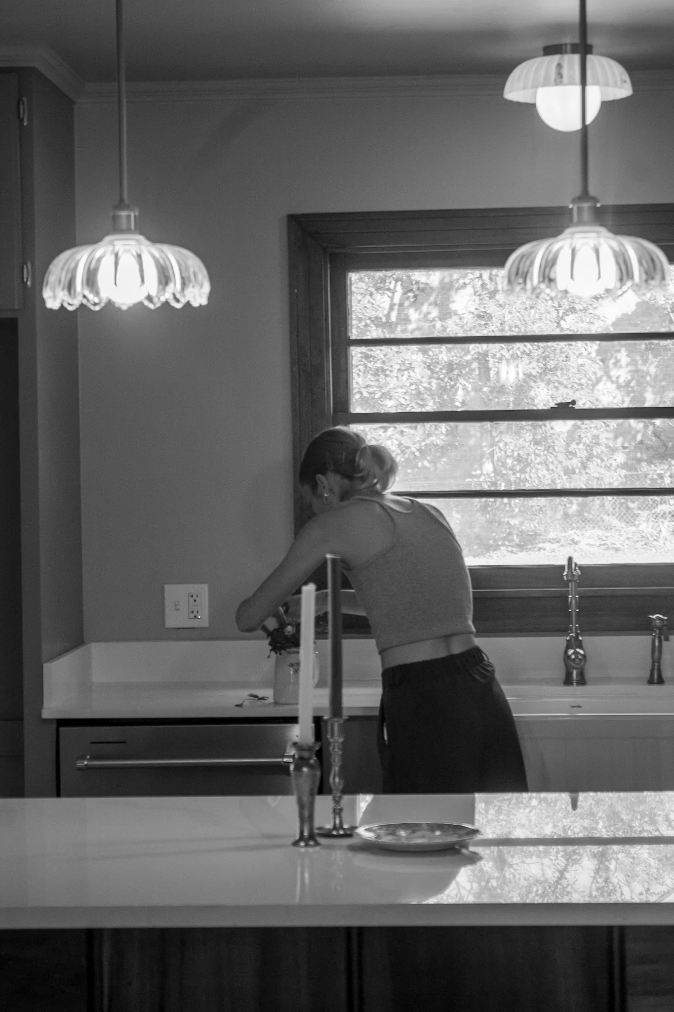 Black and white photo of designer Shelby Youtsas staging the final kitchen at 105 Teche, with vintage-inspired brass faucet, warm wood window trim, and soft pendant lighting—capturing the final moments of a Lafayette flip designed with intention.