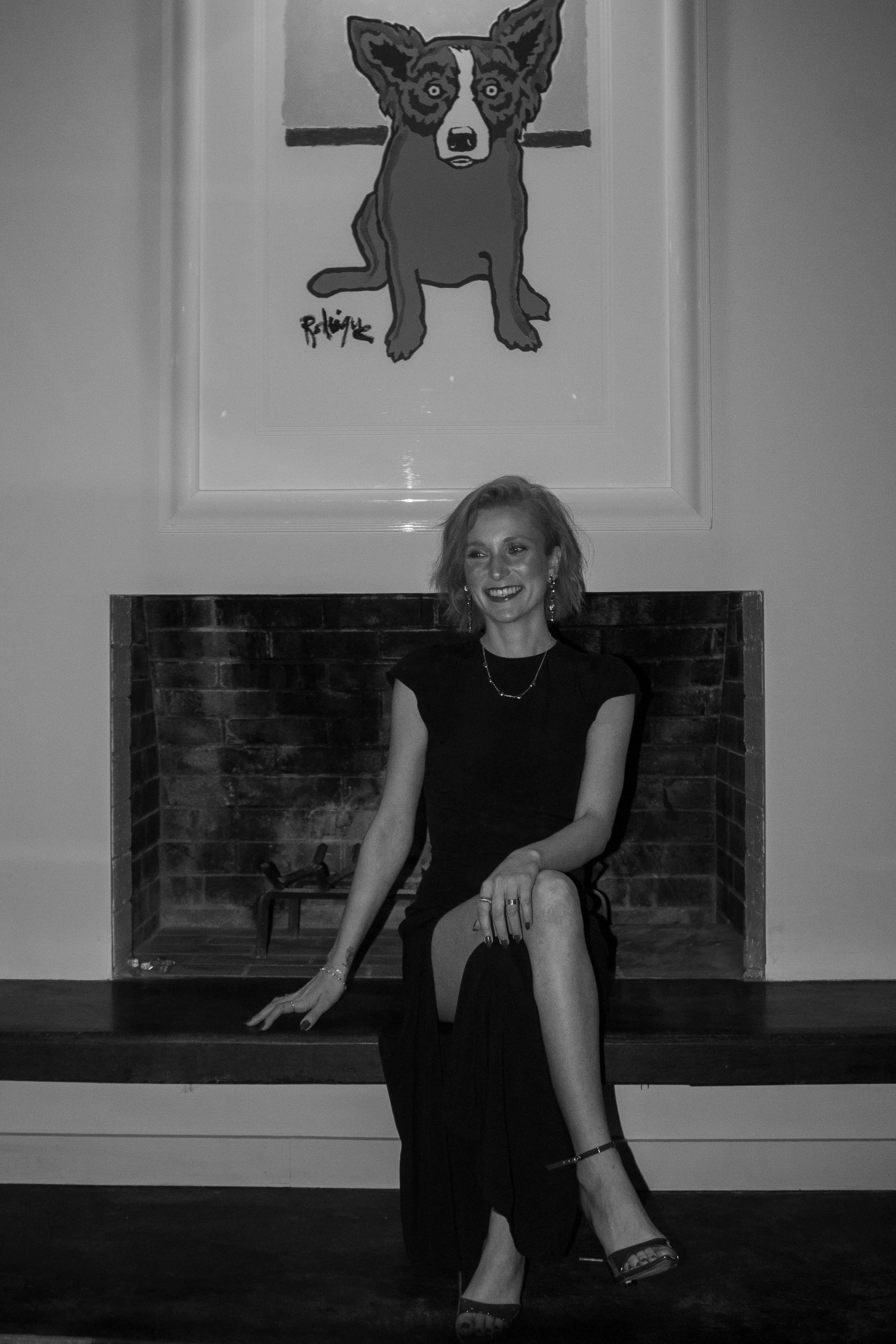 Black and white portrait of realtor Shelby Youtsas Brignac seated in front of a brick fireplace, wearing a sleek black dress, with a framed George Rodrigue blue dog painting displayed above the mantel—capturing a refined, art-forward interior moment.