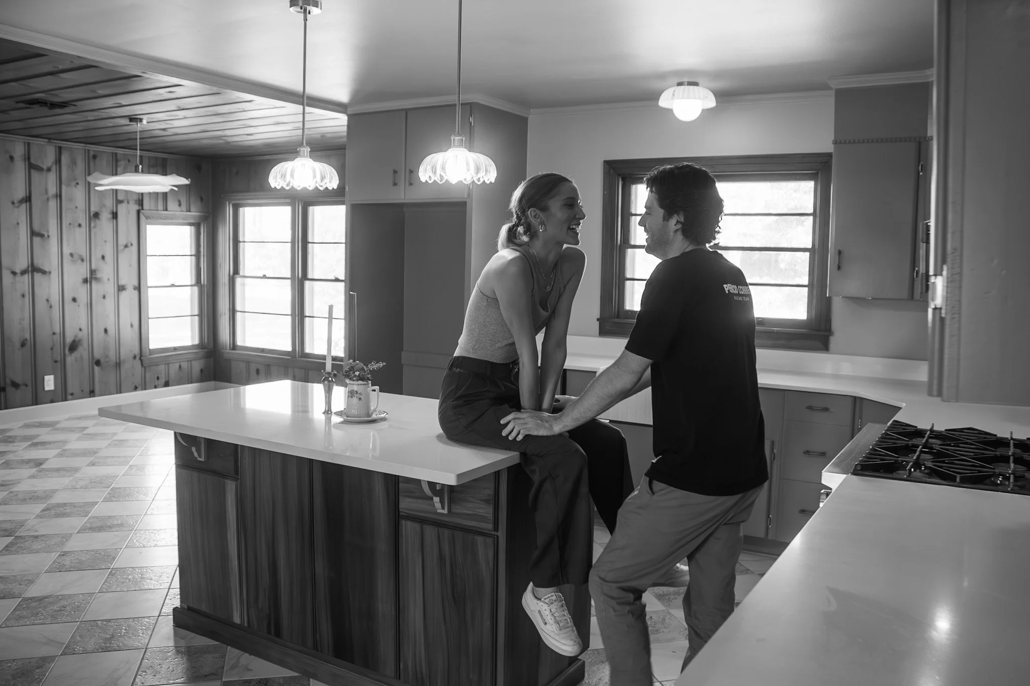 Black and white image of couple in renovated kitchen at 105 Teche in Lafayette, featuring custom walnut island, vintage-inspired lighting, and original wood paneling—capturing a candid, joyful moment that reflects the heart behind the design.