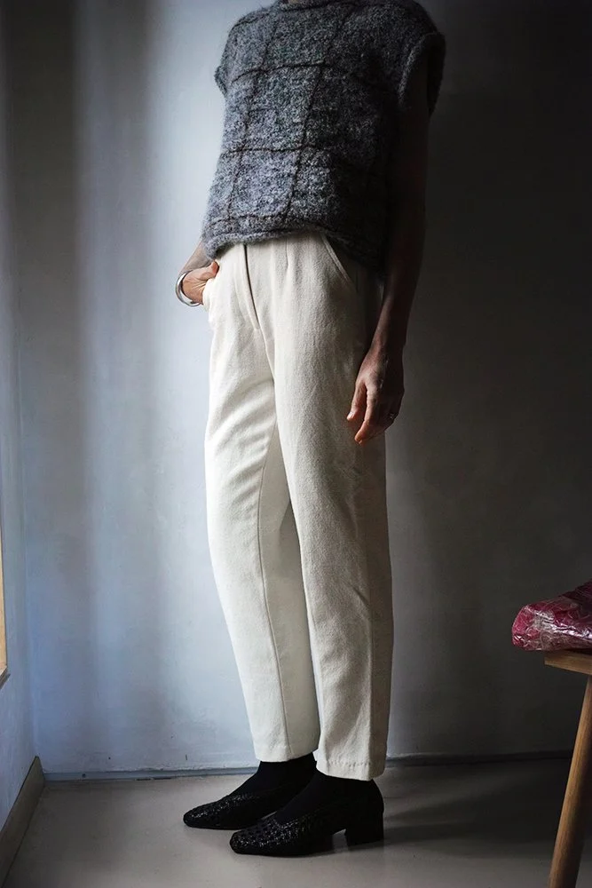 Person wearing a gray textured vest, beige high-waisted pants, and black patterned heels standing indoors near a window with a plain wall background.