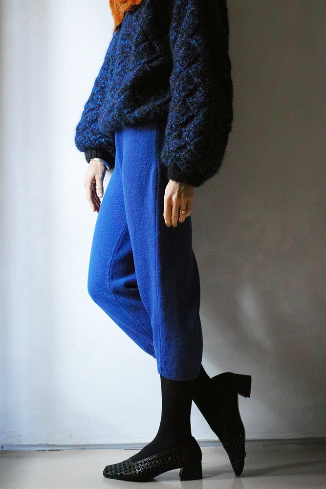 Person wearing a dark, fuzzy sweater, bright blue pants, black tights, and black heeled shoes, standing against a neutral background.