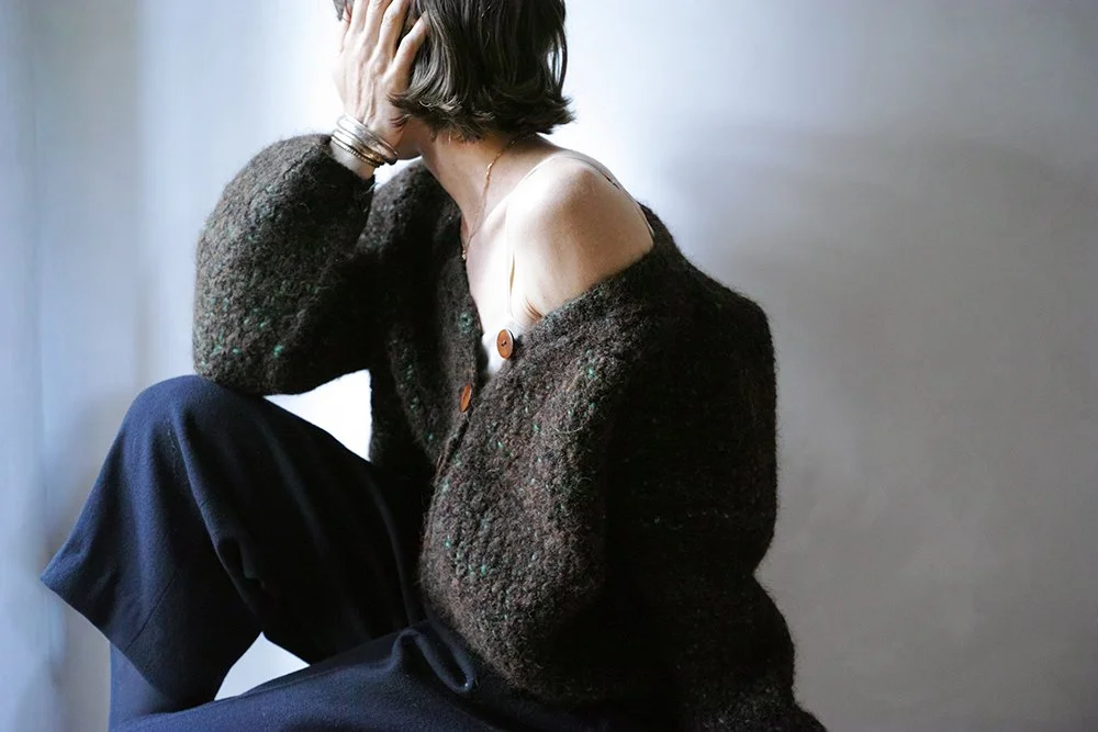 A woman with short brown hair sitting with her face covered by her hand, wearing a dark fuzzy cardigan and black wide-leg pants, against a plain background.
