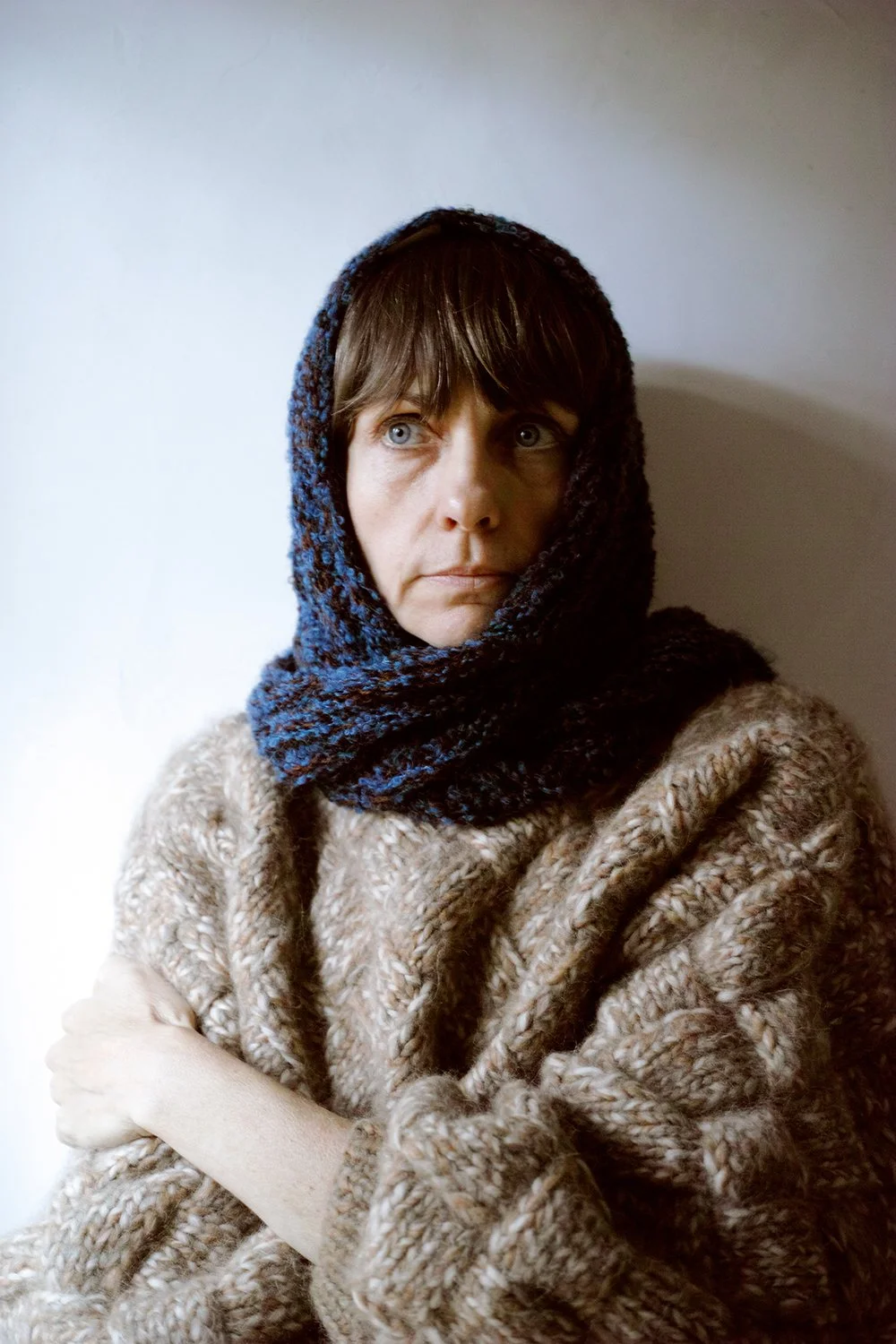 Woman with short brown hair wearing a blue knit headscarf and a thick beige sweater, looking thoughtfully to the side against a plain white wall.