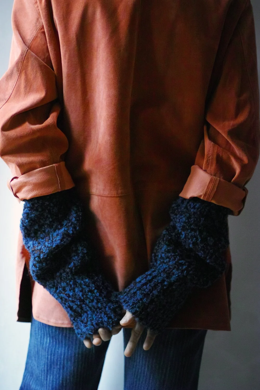 Person wearing a brown leather jacket and dark blue knit gloves, with hands clasped in front, against a neutral background.