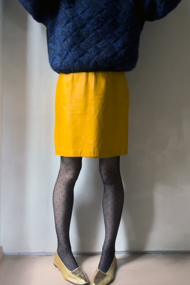 Person wearing a dark textured sweater, a yellow skirt, patterned tights, and beige shoes, standing against a plain wall.