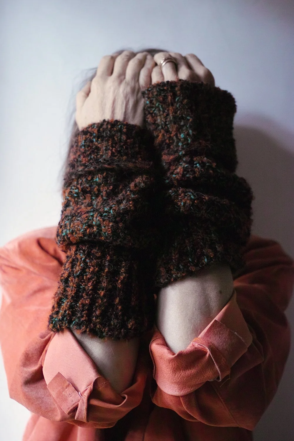 Person with hands covering face, wearing a multicolored, textured sweater and a pink jacket, standing against a plain backdrop.