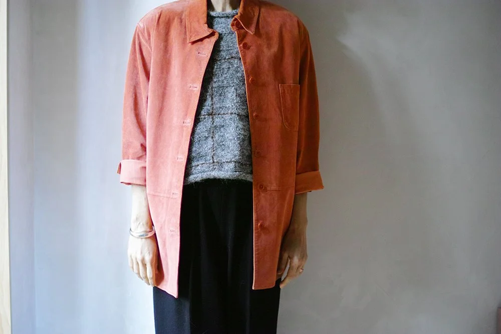 Person wearing a rust-colored jacket over a gray sweater and black pants, standing against a plain wall.