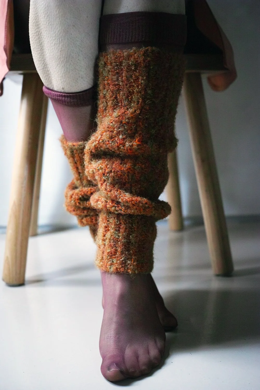 A person is wearing multicolored, fuzzy, knitted leg warmers and purple stockings while sitting on a wooden chair.