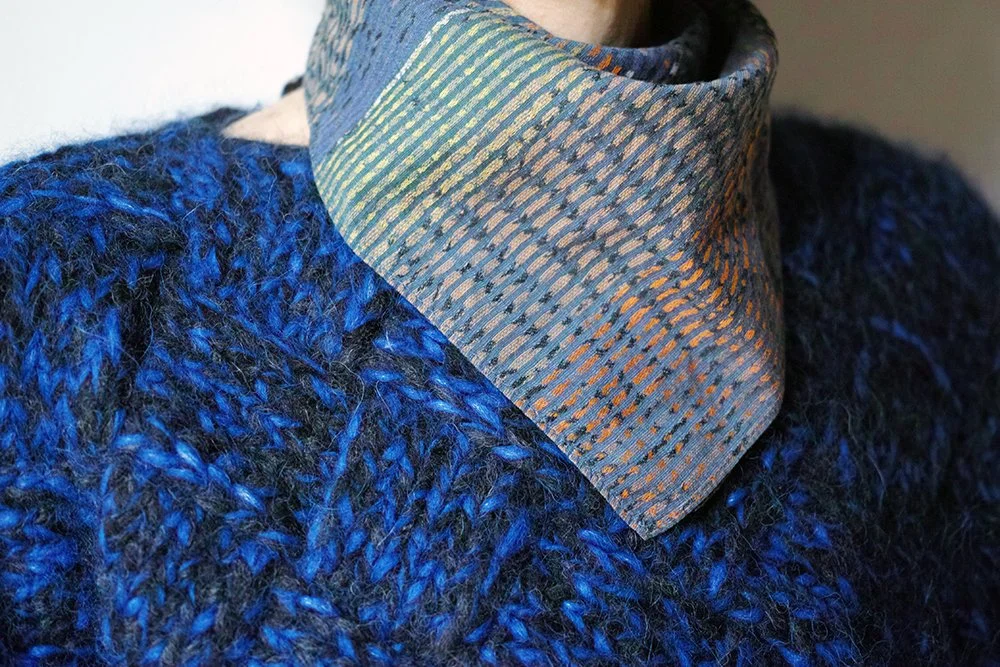 A close-up of a person wearing a blue, fuzzy, knitted sweater and a patterned fabric scarf around their neck.