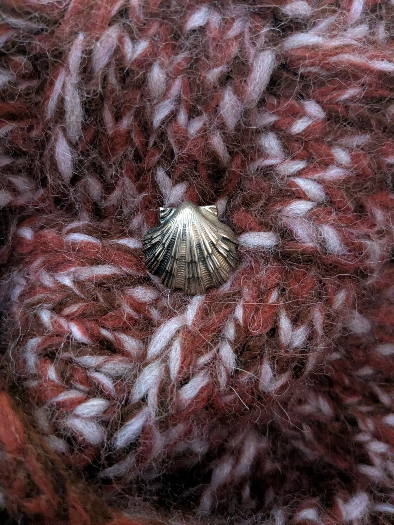 A close-up of a metallic shell-shaped pendant on a multicolored, fuzzy, knitted fabric with shades of brown, red, and white.