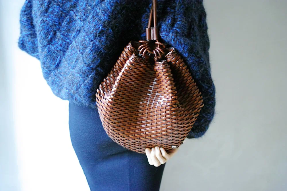 Person holding a brown woven leather handbag, wearing a blue sweater and black pants.
