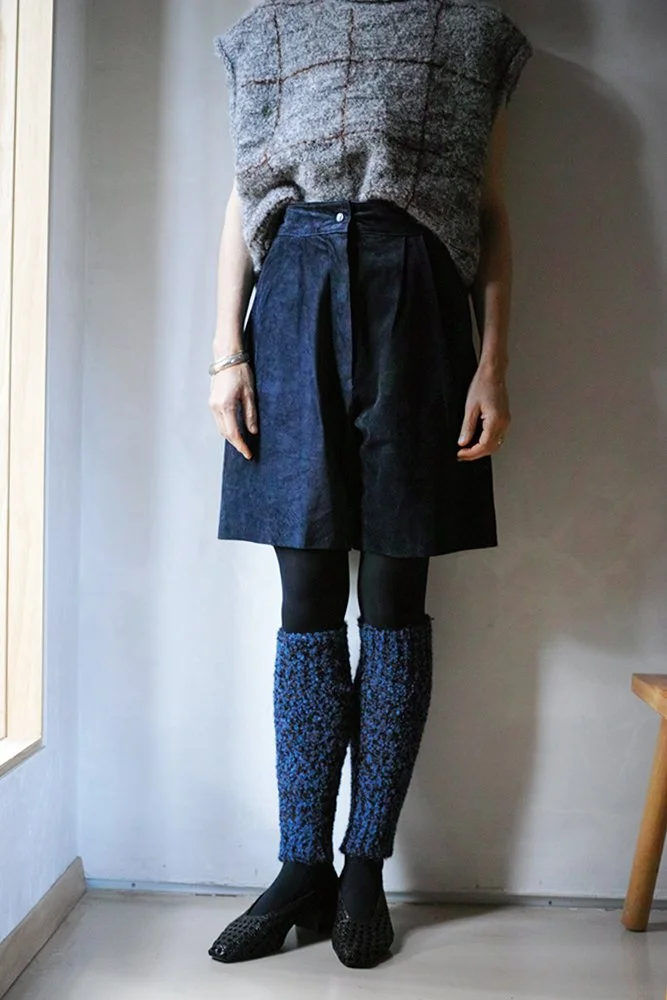 Woman standing indoors wearing a gray sleeveless top, dark knee-length wide-leg shorts, black tights, and blue patterned leg warmers with black heels.