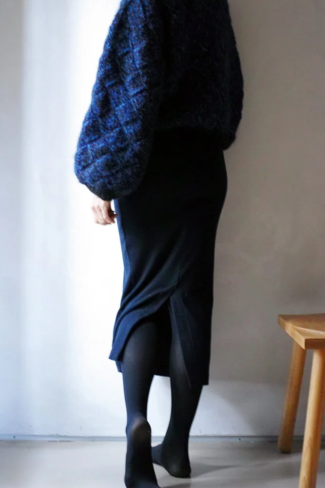 Person wearing a black skirt and black tights, standing near a gray wall and a wooden table.