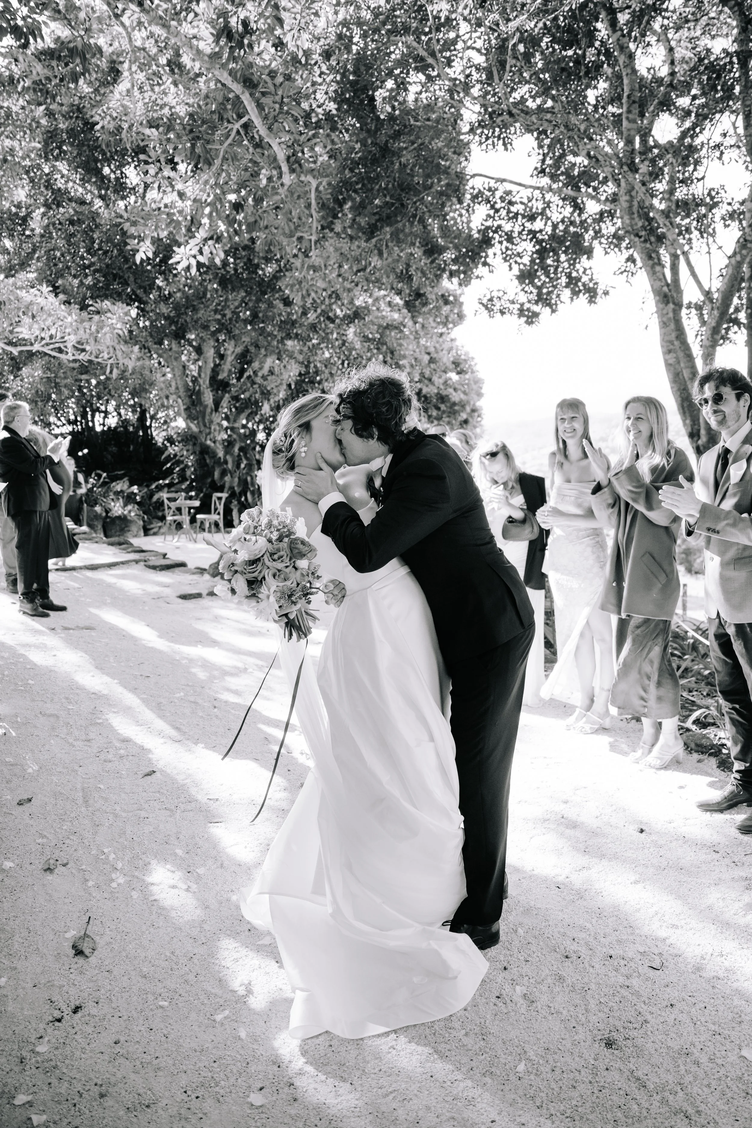A wedding couple sharing a kiss outdoors, with guests smiling and clapping in the background, surrounded by trees.