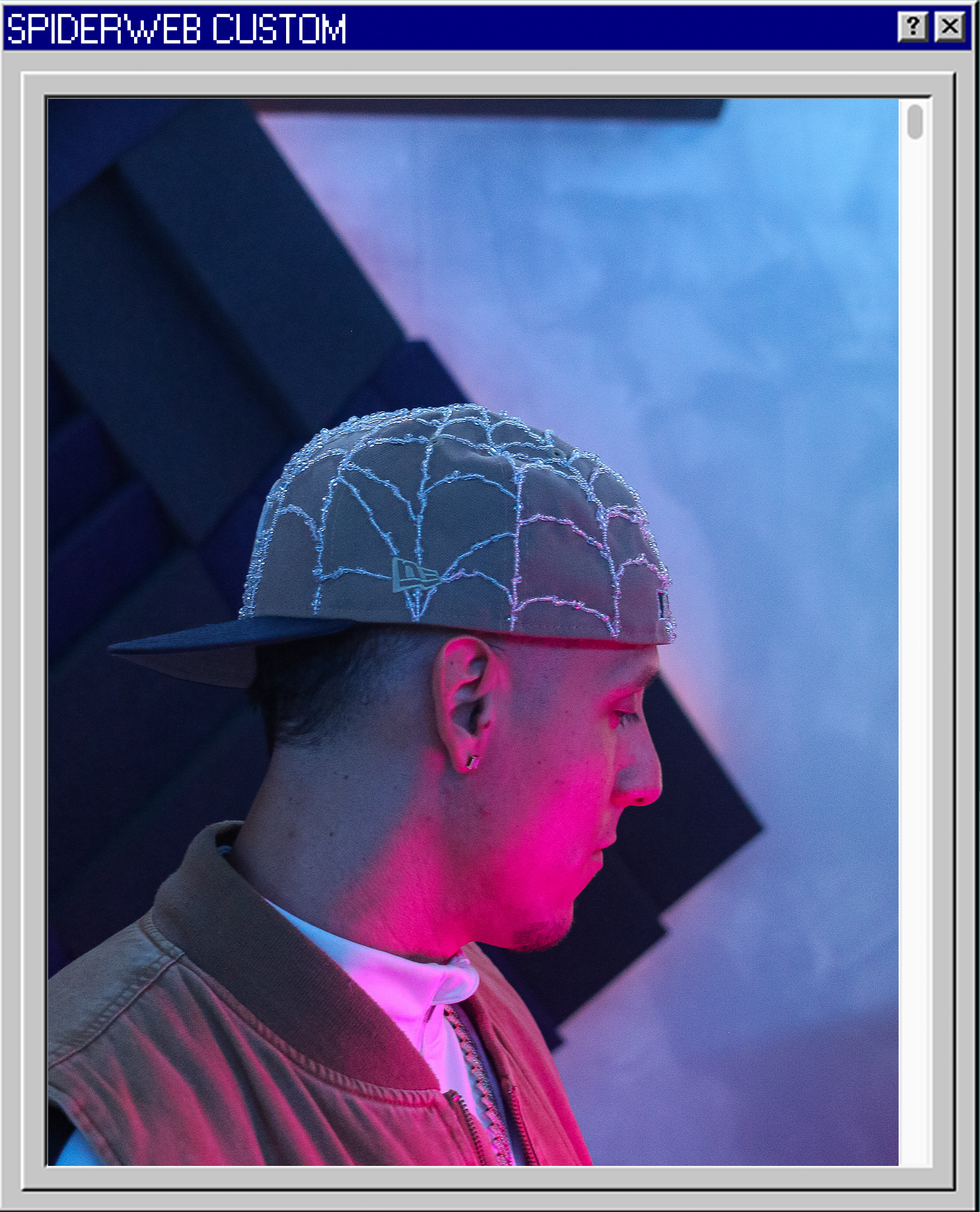 Side profile of a young man wearing a decorated baseball cap, beige jacket, and earrings, illuminated by pink and blue neon lights against a geometric background.