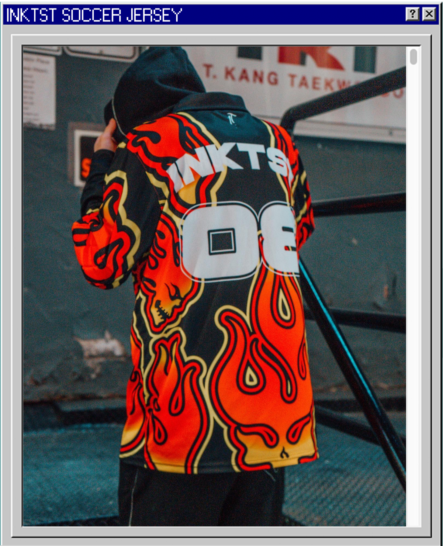 A person wearing a colorful soccer jersey with flames and skull designs, holding a hoodie up to their face, in an indoor setting with a banner in the background.