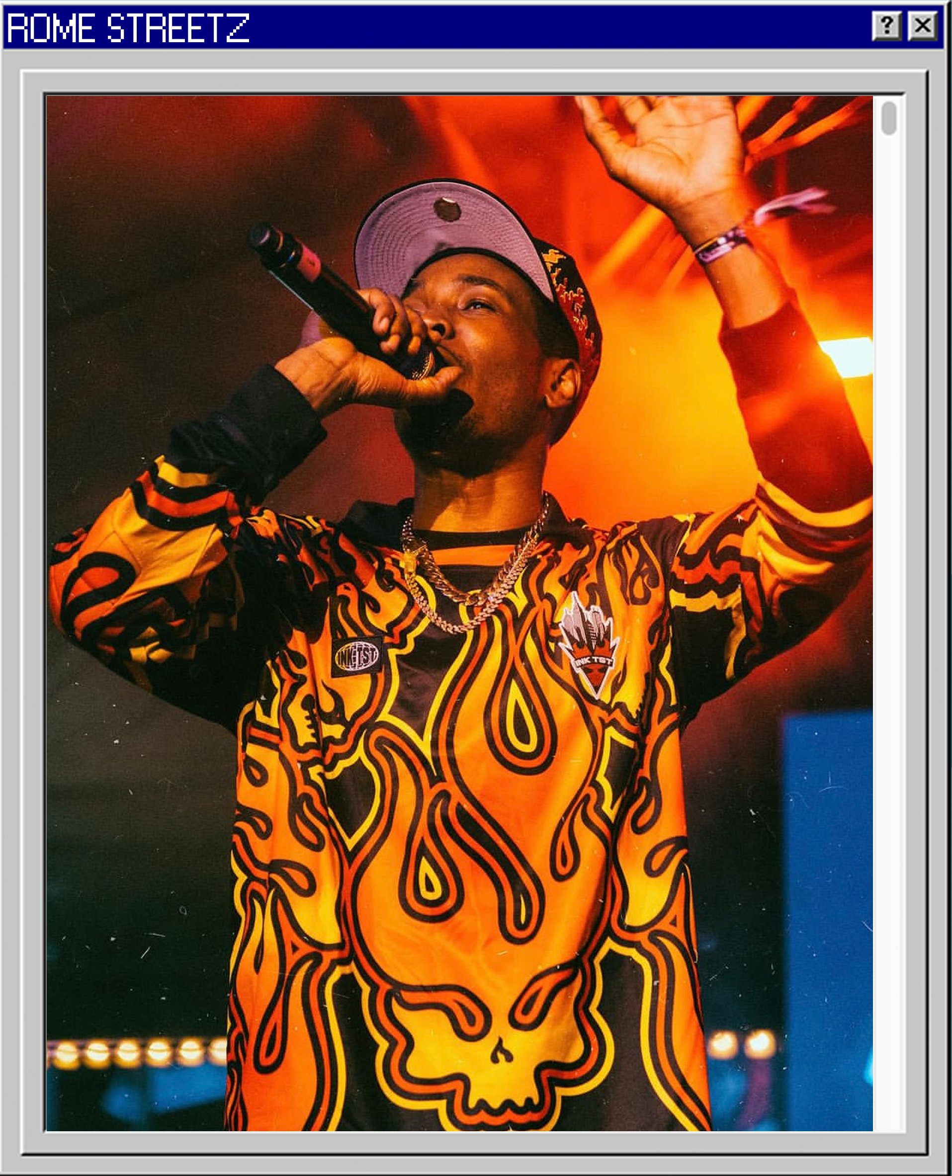 A male performer on stage holding a microphone and wearing a colorful hoodie with a skull design, a baseball cap, and jewelry, with stage lighting in the background.