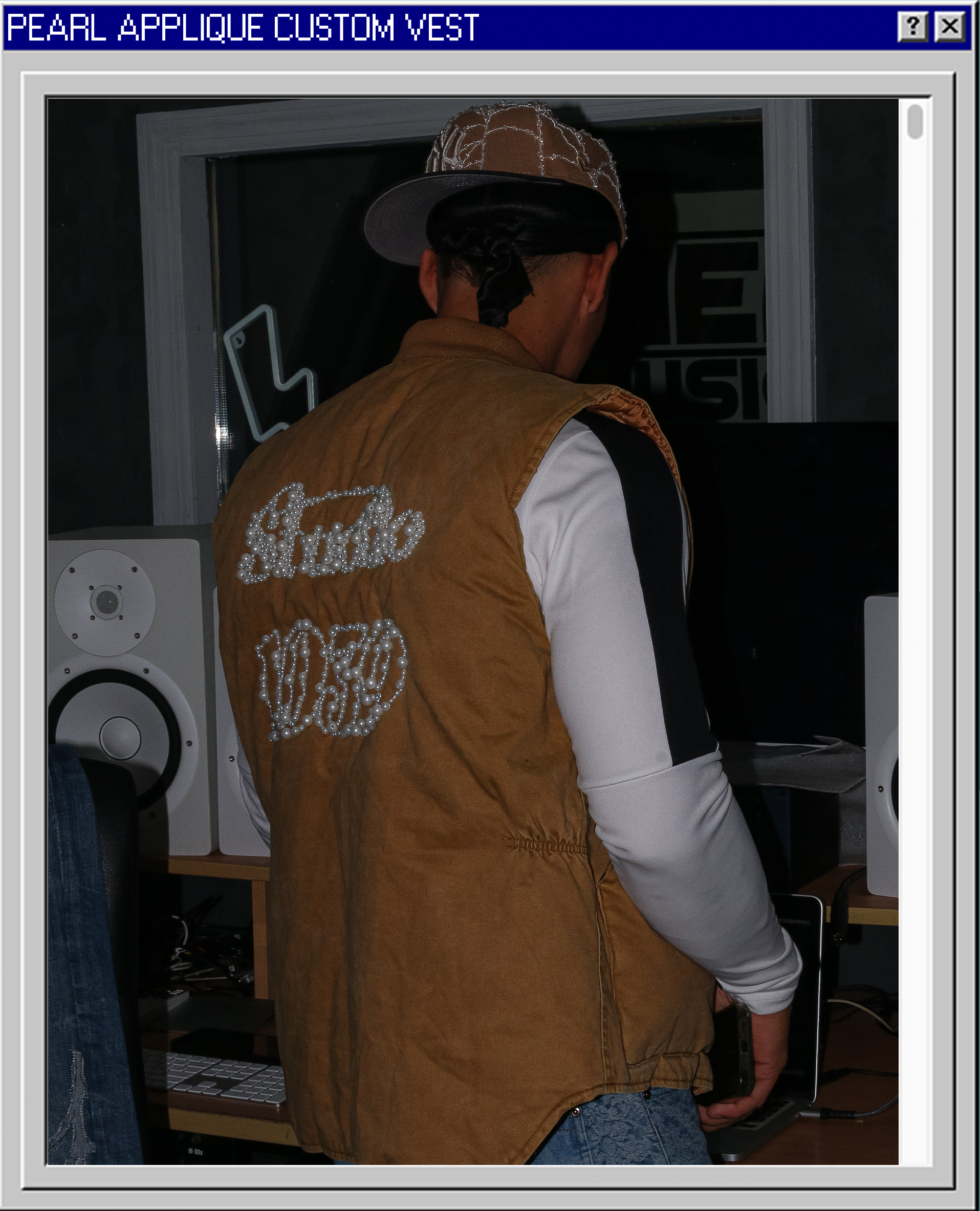 Person standing in front of a computer in a recording studio, wearing a brown vest with pearl embellishments spelling 'Luxury' and '1990,' a beige cap, and a long-sleeve white shirt with black accents.