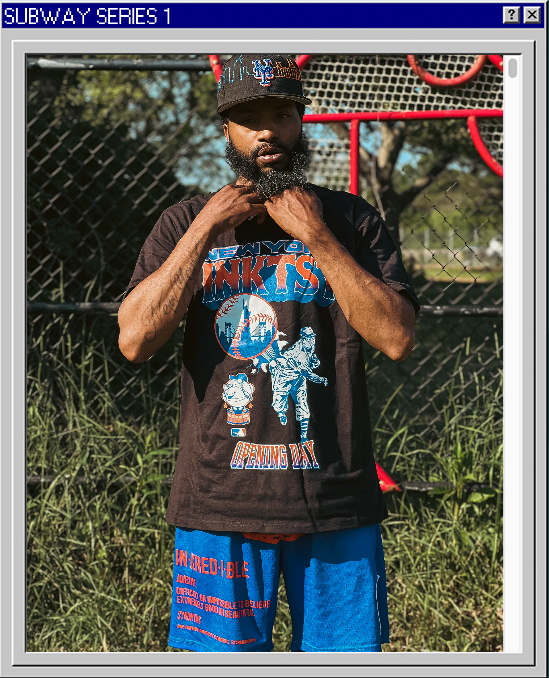 A man with a beard and mustache wearing a New York Mets cap and T-shirt standing in front of a chain-link fence and playground equipment outside.