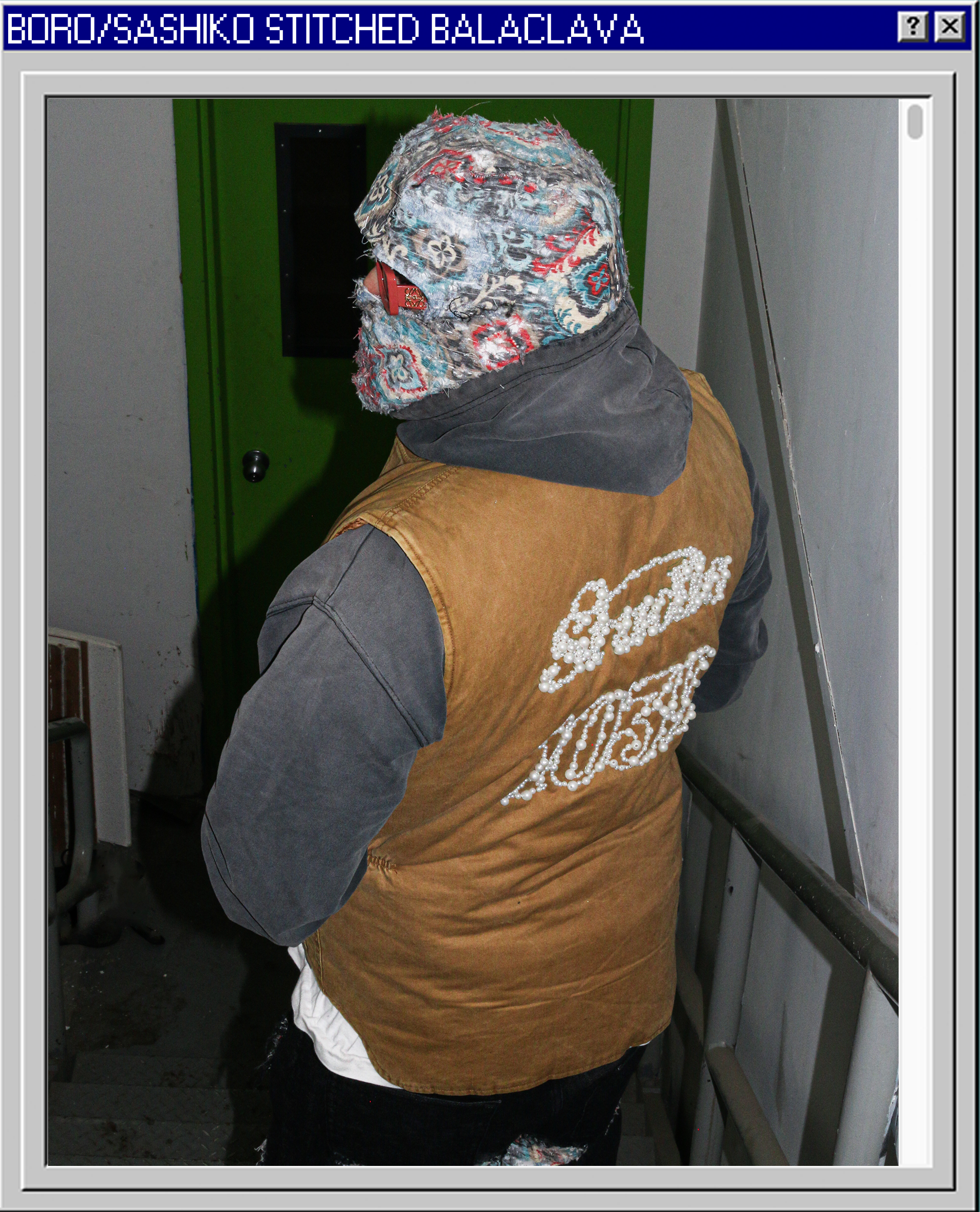 Person wearing a colorful bandana, a brown and gray jacket with white bead lettering, standing on stairs near a green door.