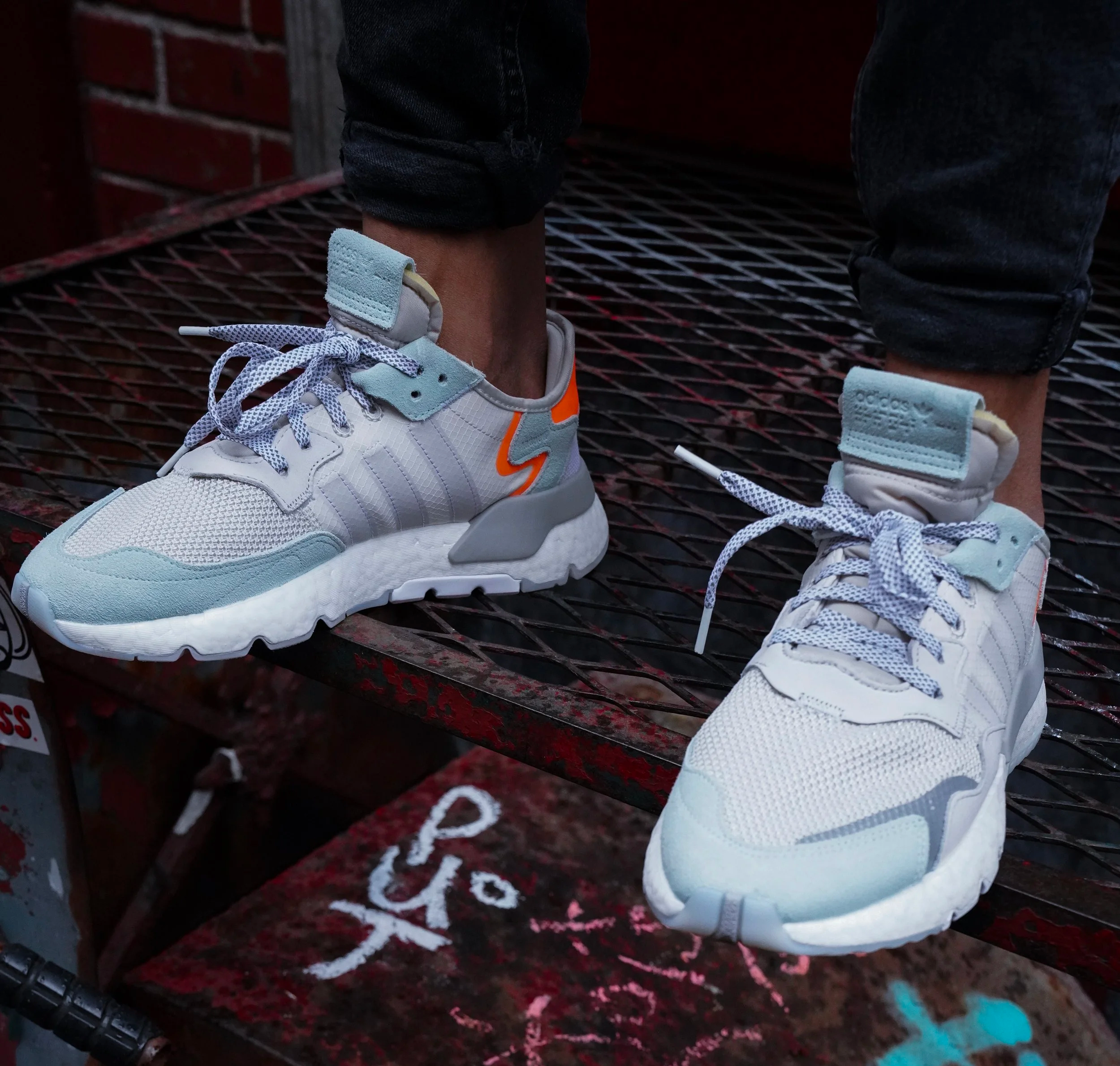 A person wearing white and light blue sneakers with reflective accents, standing on a rusty metal surface with graffiti. The person is dressed in rolled-up black jeans.