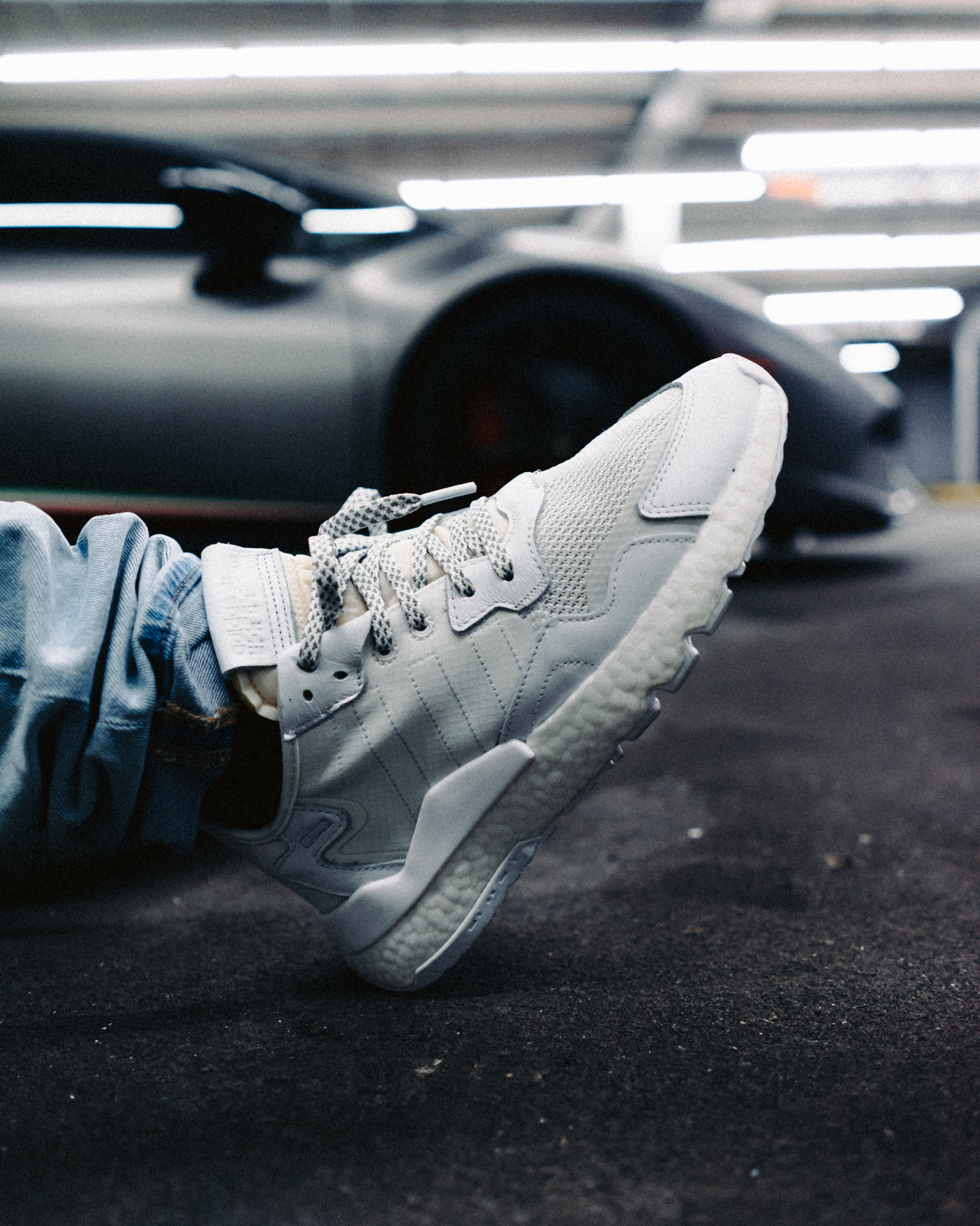 A person wearing white sneakers and jeans lying on the ground next to a black sports car in an underground parking garage.