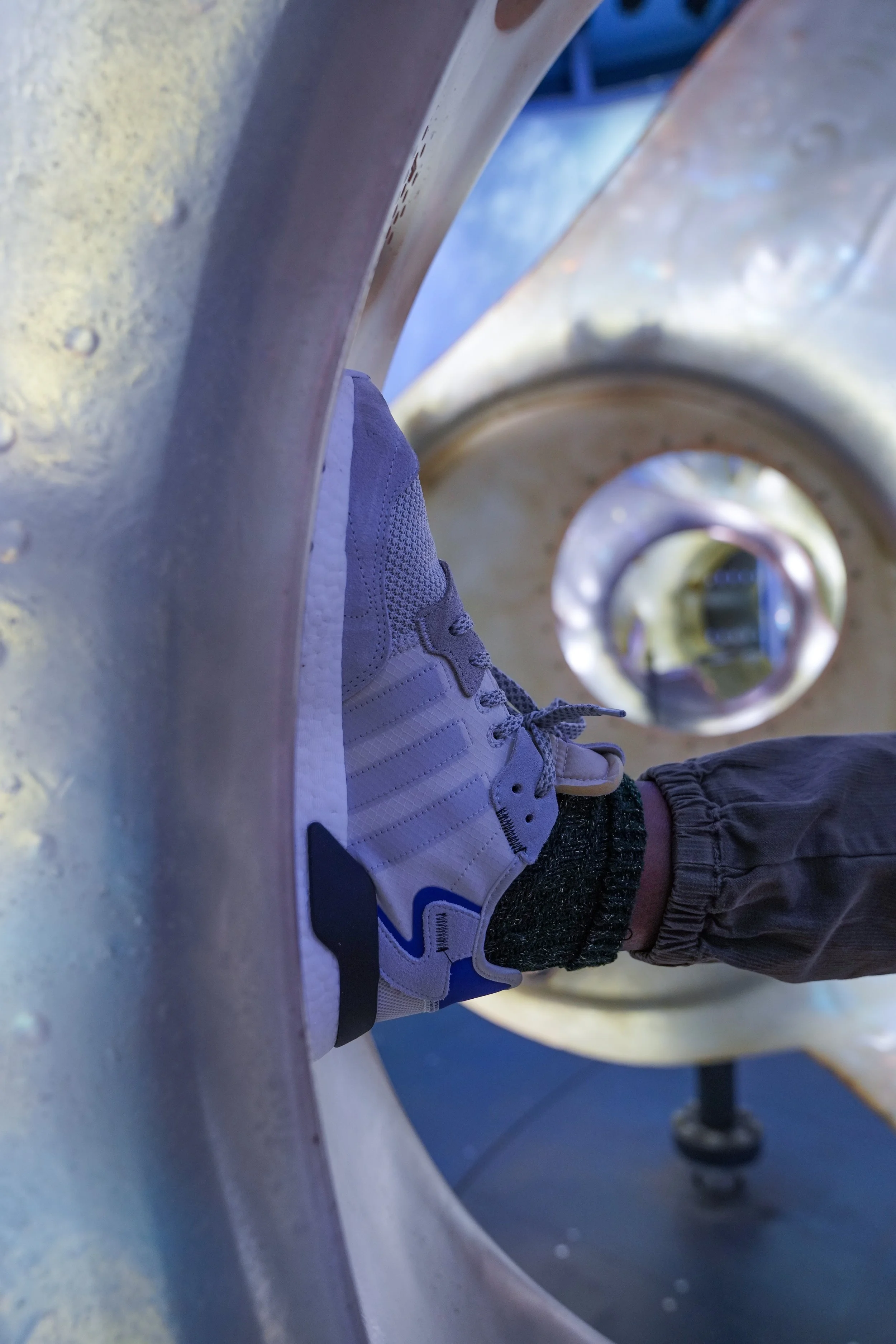 A person's foot in a white and gray sneaker resting on the edge of a metallic structure, viewed through a circular opening in the structure.
