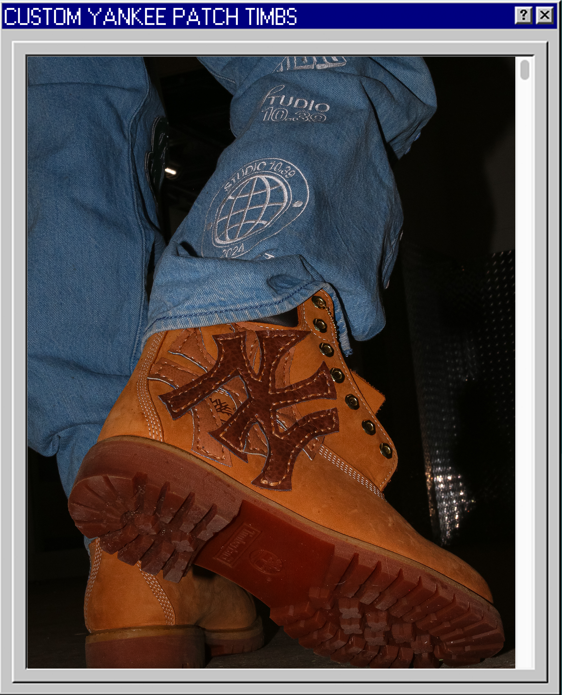 Close-up of a brown Leatherface-style boot with a large black cross design on the side. The boot is on a person's foot, with the ground visible. The person is wearing blue jeans with patches and embroidery.