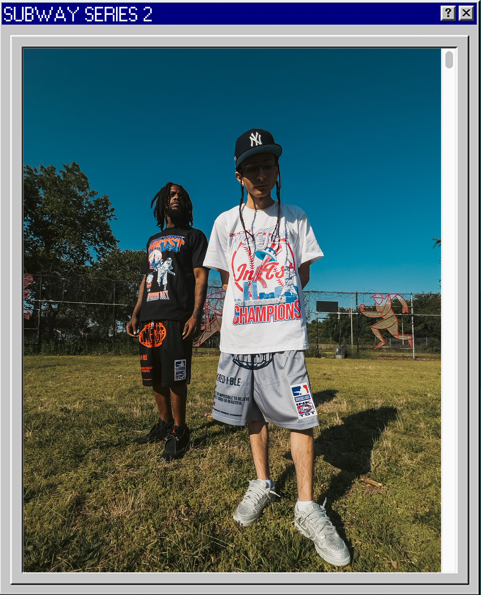 Two young men standing on a grassy field with a clear blue sky and a chain-link fence in the background. One is wearing a black T-shirt with graphic design, black shorts, and black sneakers. The other is wearing a white graphic T-shirt, gray shorts, 