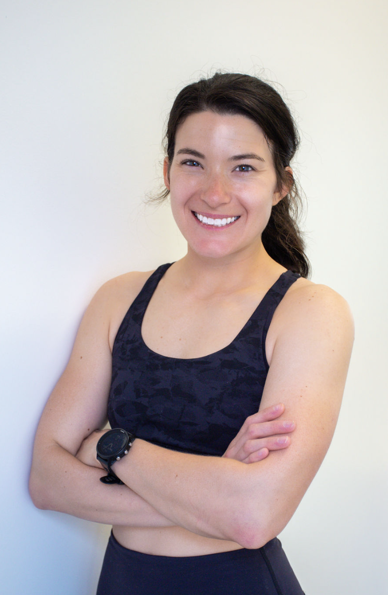 A woman smiling with her arms crossed, wearing a black athletic tank top and a watch, standing against a plain white wall.