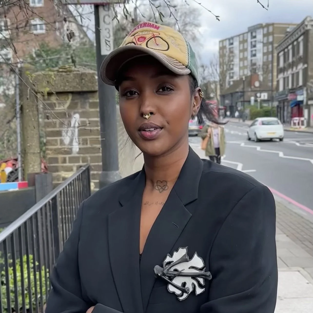 A person wearing a black blazer with a decorative pin, a cap with a bicycle design, and a septum piercing, standing on an urban street with buildings in the background.