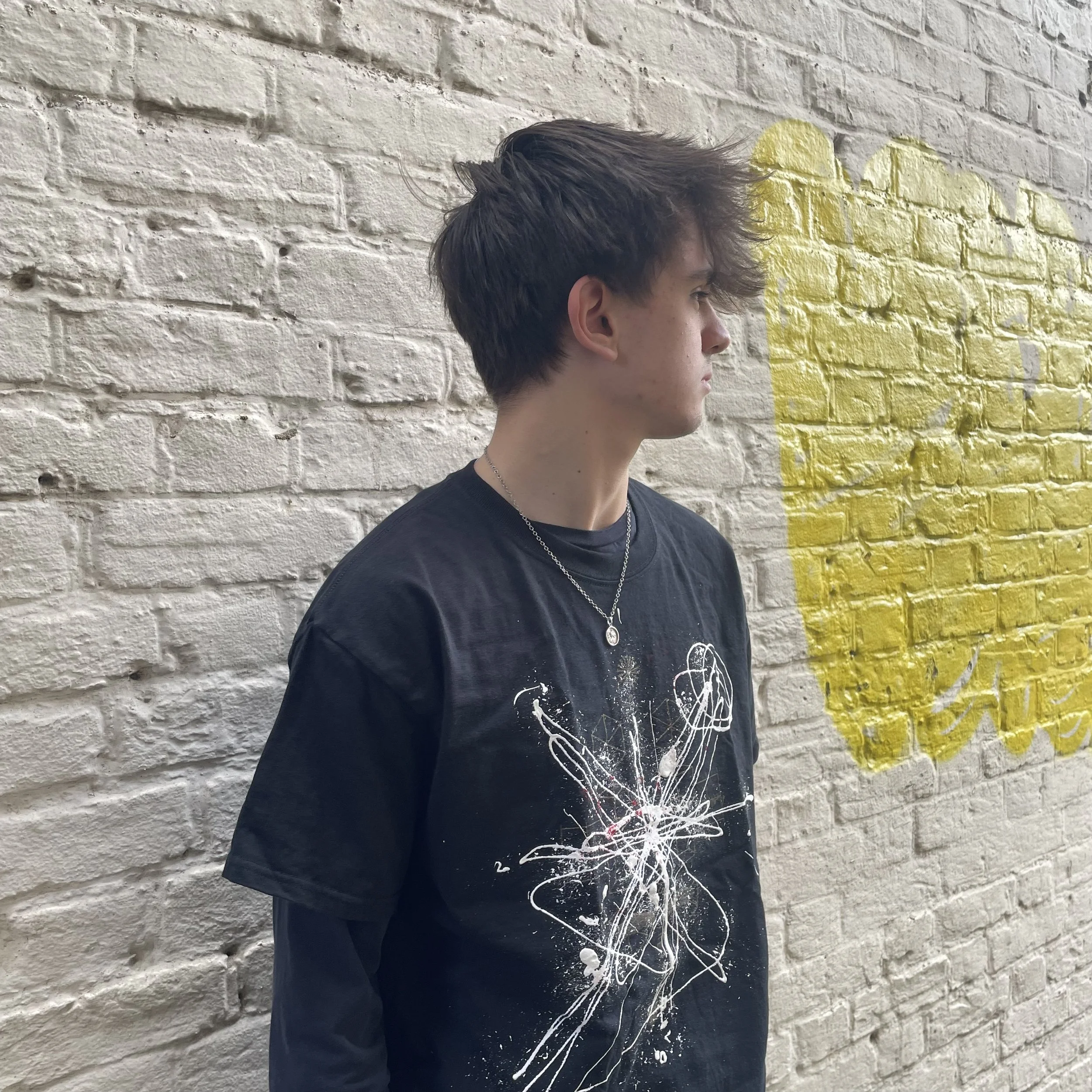 Person with short hair and a black shirt with abstract white design, standing against a white brick wall with yellow graffiti.
