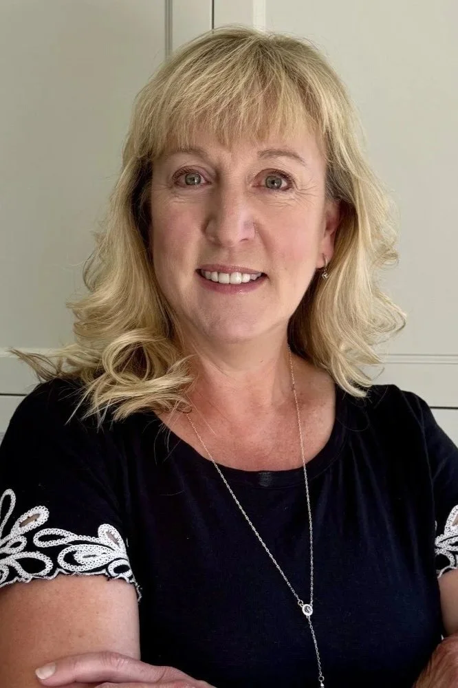 A woman with blonde hair smiling at the camera, wearing a black top with white lace detail on the sleeves and a silver necklace, standing indoors.